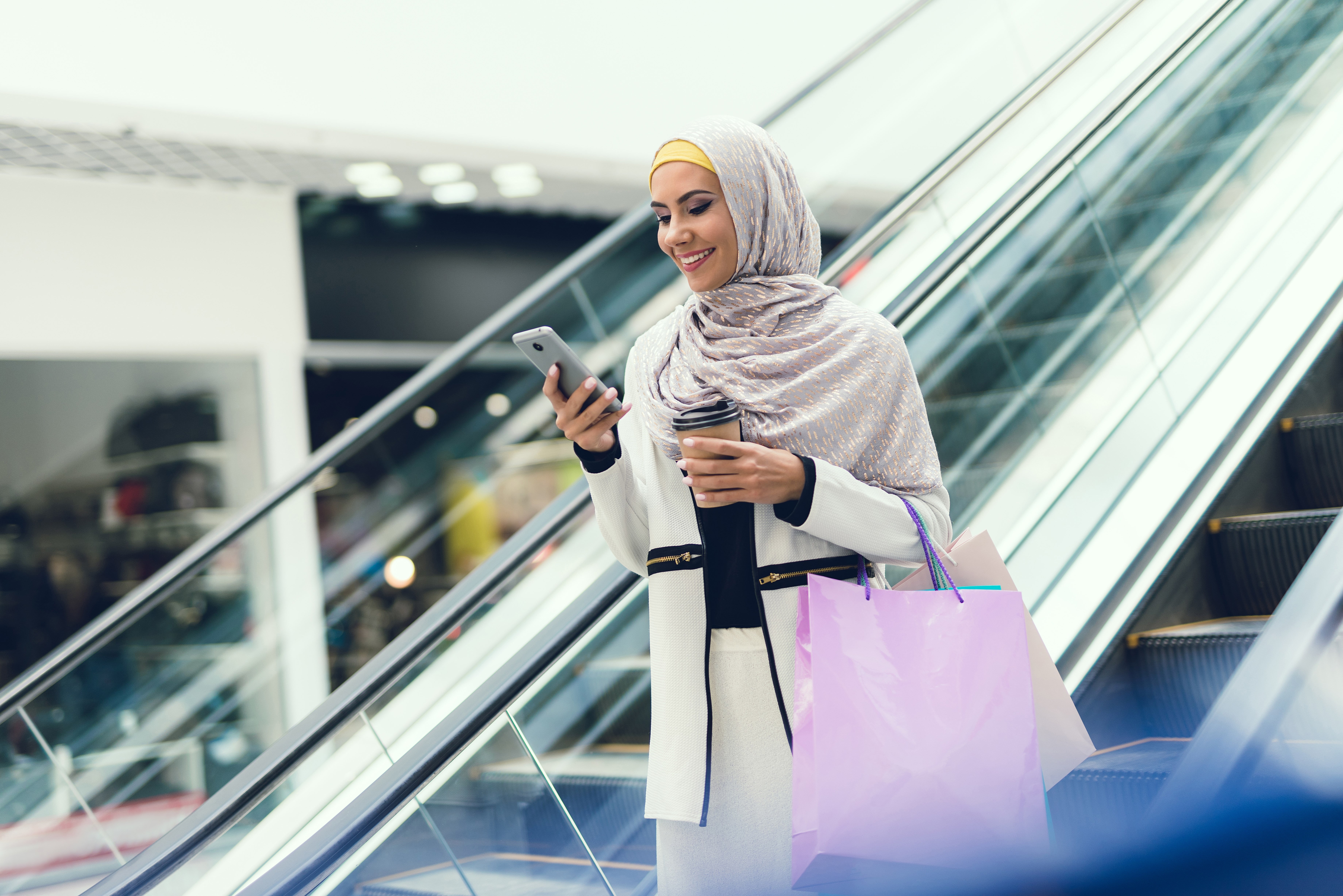 Woman smiling while holding phone in hand. Woman with packages in hands walks through the mall.