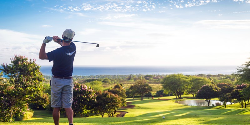 Man hitting golf ball down hill towards ocean and horizon