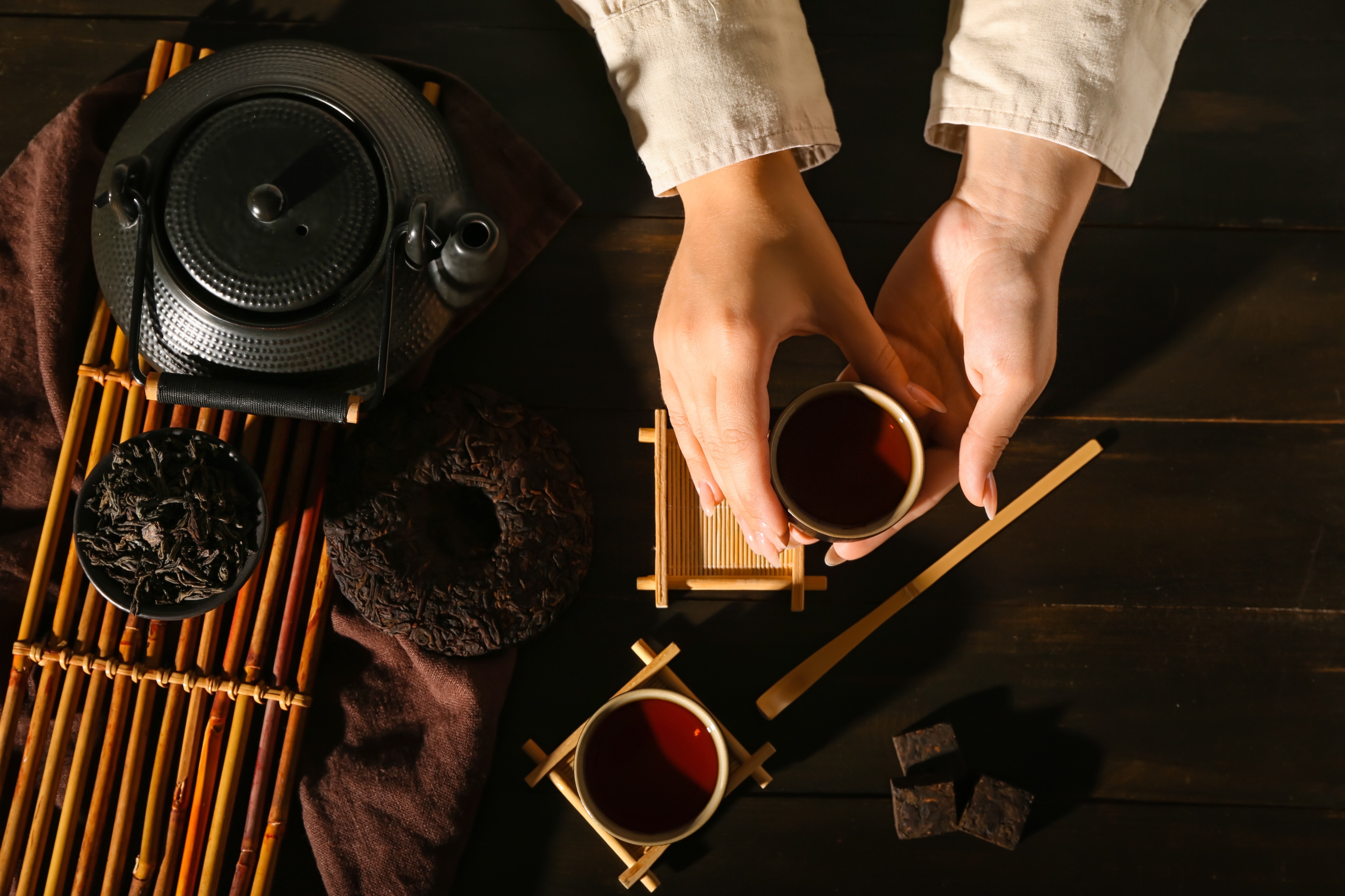 Female hands with cup of hot puer tea and dry pressed leaves on dark wooden table