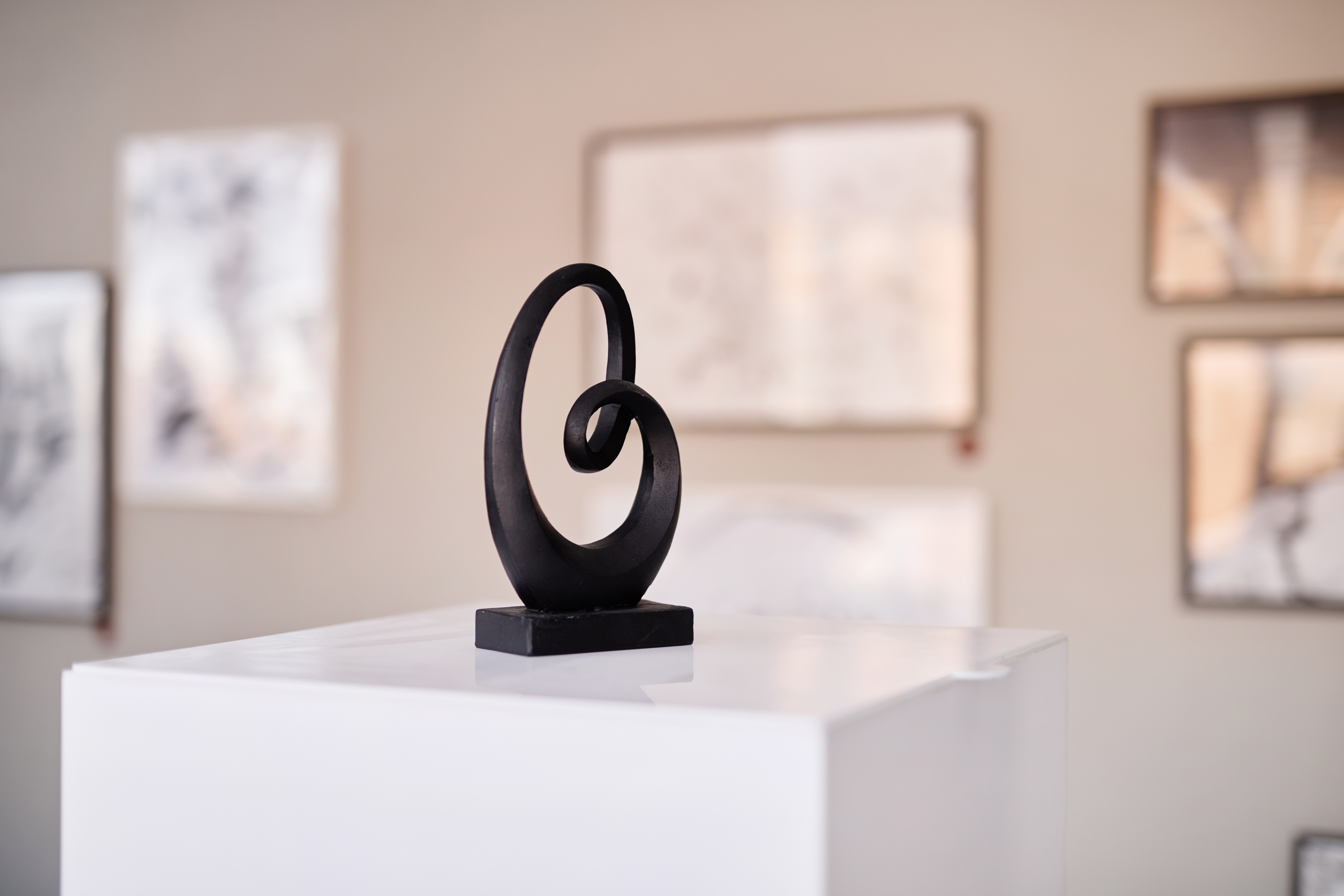 Abstract black sculpture on pedestal, blurred artworks behind emphasizing form