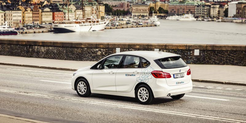 En vit bilpoolsbil från DriveNow med siluetten av Gamla Stan Stockholm bakom En vit bilpoolsbil från DriveNow med siluetten av Gamla Stan Stockholm bakom